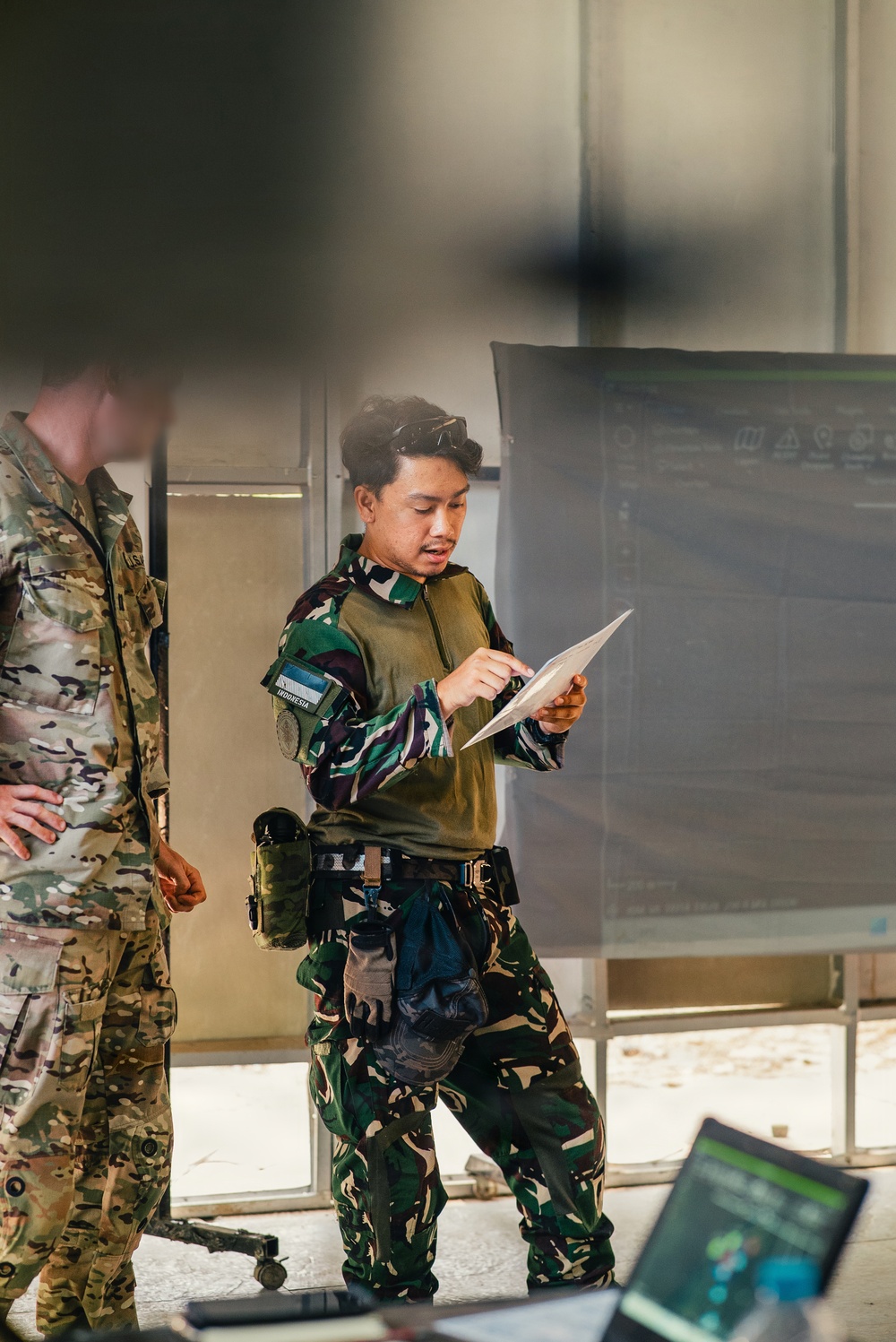 Two military personnel are visible in what appears to be an indoor operations or briefing area. The central figure, wearing Indonesian Armed Forces (TNI) camouflage uniform with an 'INDONESIA' name tape and flag patch, is reviewing a document or map. A second soldier in MultiCam/OCP pattern uniform, likely US military, is partially visible on the left.