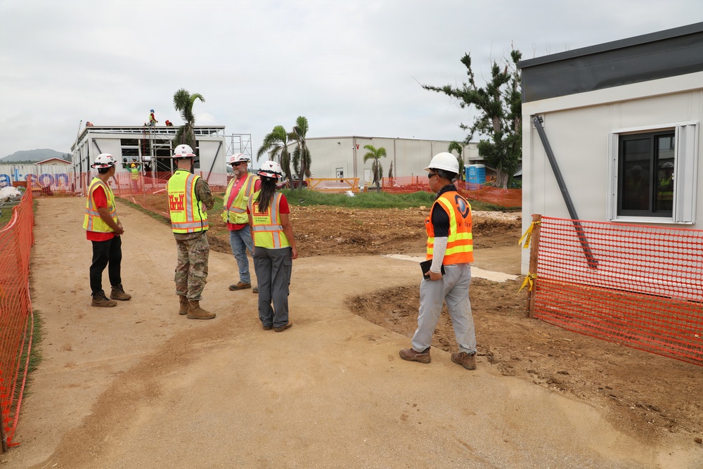A group of five individuals are gathered at an active construction site, wearing high-visibility safety vests and hard hats. The site features partially constructed modular or prefabricated buildings, orange safety fencing, and cleared dirt pathways. One individual appears to be in military camouflage uniform among the construction personnel.