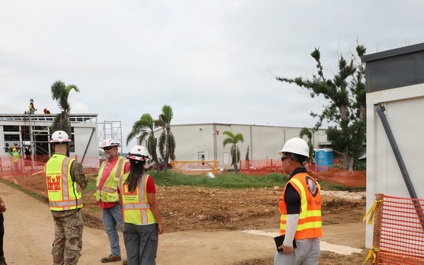 A group of five individuals are gathered at an active construction site, wearing high-visibility safety vests and hard hats. The site features partially constructed modular or prefabricated buildings, orange safety fencing, and cleared dirt pathways. One individual appears to be in military camouflage uniform among the construction personnel.