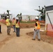 A group of five individuals are gathered at an active construction site, wearing high-visibility safety vests and hard hats. The site features partially constructed modular or prefabricated buildings, orange safety fencing, and cleared dirt pathways. One individual appears to be in military camouflage uniform among the construction personnel.