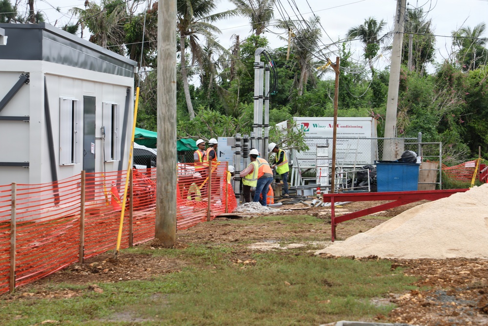 An active outdoor construction site featuring several workers in high-visibility safety vests and hard hats working near equipment and materials. The site is bordered by orange safety fencing and includes a modular site office building on the left. The background shows palm trees with storm-damaged or stripped foliage, suggesting a tropical location possibly recovering from a weather event.