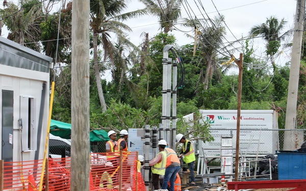 An active outdoor construction site featuring several workers in high-visibility safety vests and hard hats working near equipment and materials. The site is bordered by orange safety fencing and includes a modular site office building on the left. The background shows palm trees with storm-damaged or stripped foliage, suggesting a tropical location possibly recovering from a weather event.
