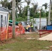 An active outdoor construction site featuring several workers in high-visibility safety vests and hard hats working near equipment and materials. The site is bordered by orange safety fencing and includes a modular site office building on the left. The background shows palm trees with storm-damaged or stripped foliage, suggesting a tropical location possibly recovering from a weather event.