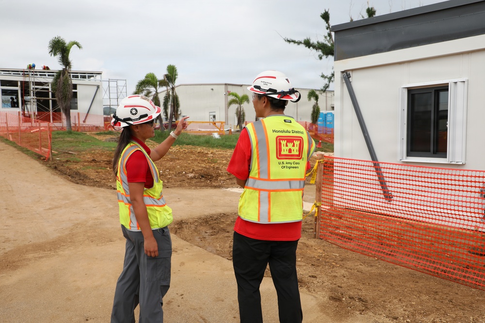 Two construction site inspectors wearing U.S. Army Corps of Engineers safety vests and white hard hats are conducting a site walkthrough. One person is pointing toward a structure or area of interest while the other observes. The site appears to be an active construction zone with temporary buildings, orange safety fencing, and palm trees visible in the background.