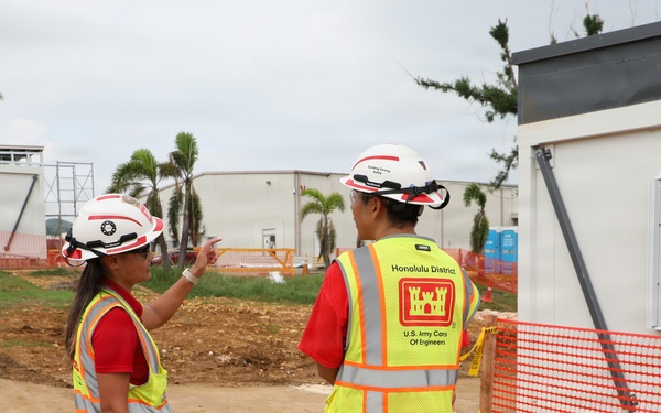 Two construction site inspectors wearing U.S. Army Corps of Engineers safety vests and white hard hats are conducting a site walkthrough. One person is pointing toward a structure or area of interest while the other observes. The site appears to be an active construction zone with temporary buildings, orange safety fencing, and palm trees visible in the background.