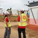 Two construction site inspectors wearing U.S. Army Corps of Engineers safety vests and white hard hats are conducting a site walkthrough. One person is pointing toward a structure or area of interest while the other observes. The site appears to be an active construction zone with temporary buildings, orange safety fencing, and palm trees visible in the background.