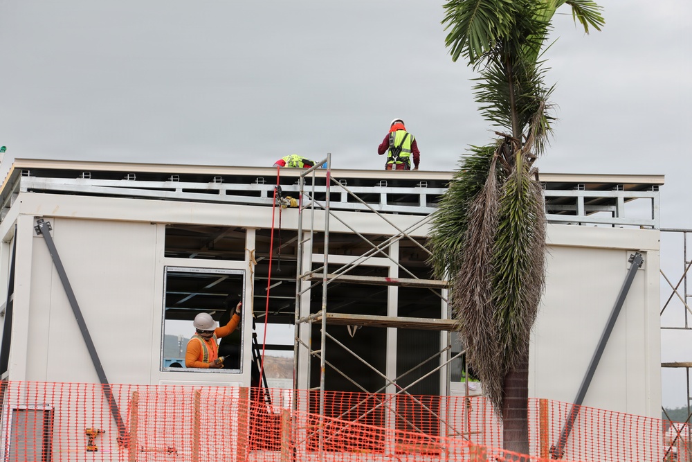 A construction site showing workers actively building or renovating a two-story structure. At least two workers are visible — one on the rooftop wearing a yellow high-visibility vest and red hard hat, and another at ground/lower level wearing an orange vest and white hard hat. Safety equipment including harnesses, orange safety fencing, and scaffolding are prominently visible.