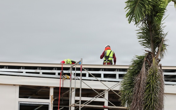 A construction site showing workers actively building or renovating a two-story structure. At least two workers are visible — one on the rooftop wearing a yellow high-visibility vest and red hard hat, and another at ground/lower level wearing an orange vest and white hard hat. Safety equipment including harnesses, orange safety fencing, and scaffolding are prominently visible.