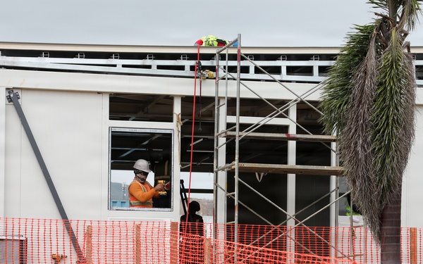 A construction site showing a modular or prefabricated building under assembly. A worker in high-visibility orange clothing and a white hard hat is visible working inside or near a window opening. The site is secured with orange plastic safety mesh fencing along the perimeter.