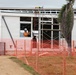 A construction site showing a modular or prefabricated building under assembly. A worker in high-visibility orange clothing and a white hard hat is visible working inside or near a window opening. The site is secured with orange plastic safety mesh fencing along the perimeter.
