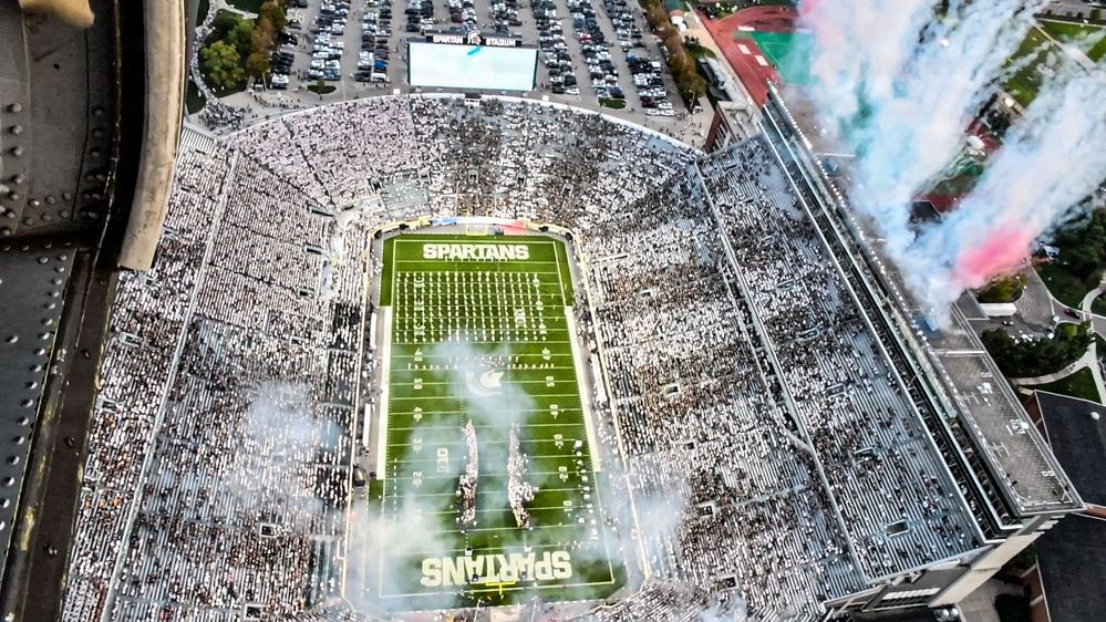 An aerial view of a packed college football stadium identified as 'Spartans' stadium, likely Spartan Stadium at Michigan State University. The stadium is filled to capacity with fans dressed predominantly in white, creating a whiteout effect. Pre-game or halftime festivities are underway, with colorful pyrotechnic smoke effects visible on the field and from the stadium structure.