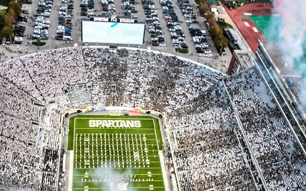 An aerial view of a packed college football stadium identified as 'Spartans' stadium, likely Spartan Stadium at Michigan State University. The stadium is filled to capacity with fans dressed predominantly in white, creating a whiteout effect. Pre-game or halftime festivities are underway, with colorful pyrotechnic smoke effects visible on the field and from the stadium structure.