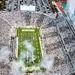 An aerial view of a packed college football stadium identified as 'Spartans' stadium, likely Spartan Stadium at Michigan State University. The stadium is filled to capacity with fans dressed predominantly in white, creating a whiteout effect. Pre-game or halftime festivities are underway, with colorful pyrotechnic smoke effects visible on the field and from the stadium structure.