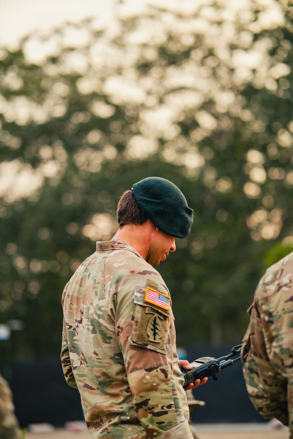 A U.S. Army Special Forces soldier wearing a green beret and MultiCam (OCP) camouflage uniform is looking downward at a piece of equipment held in his hand. He displays a U.S. flag patch on his right shoulder and what appears to be a Special Forces unit patch below it. The background is heavily blurred with trees, suggesting an outdoor setting during golden hour lighting.