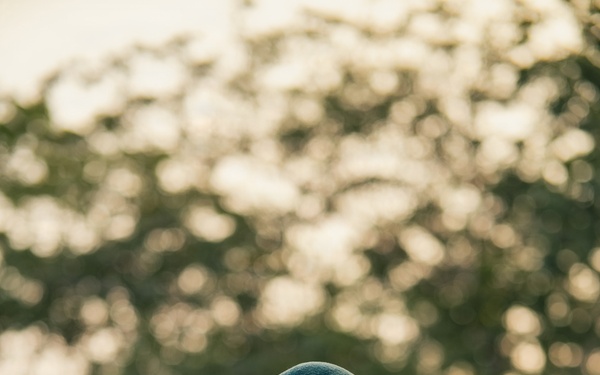 A U.S. Army Special Forces soldier wearing a green beret and MultiCam (OCP) camouflage uniform is looking downward at a piece of equipment held in his hand. He displays a U.S. flag patch on his right shoulder and what appears to be a Special Forces unit patch below it. The background is heavily blurred with trees, suggesting an outdoor setting during golden hour lighting.