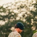 A U.S. Army Special Forces soldier wearing a green beret and MultiCam (OCP) camouflage uniform is looking downward at a piece of equipment held in his hand. He displays a U.S. flag patch on his right shoulder and what appears to be a Special Forces unit patch below it. The background is heavily blurred with trees, suggesting an outdoor setting during golden hour lighting.