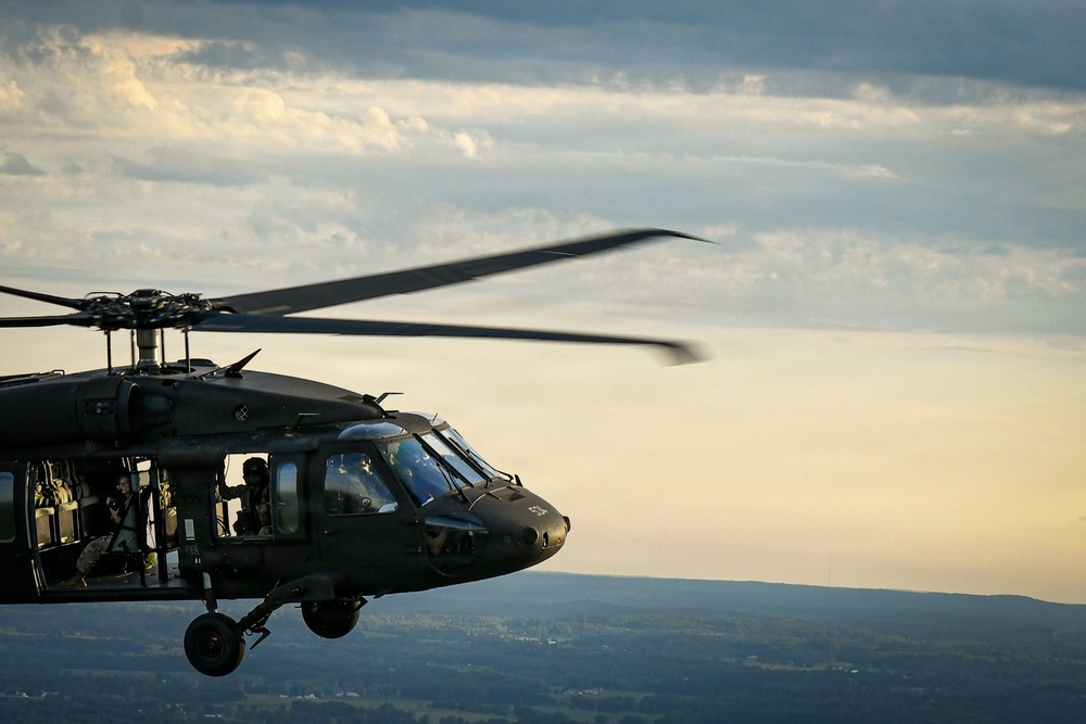 A military helicopter, identifiable as a UH-60 Black Hawk, is captured in flight against a partly cloudy sky at what appears to be dusk or golden hour. The helicopter is painted in dark olive/matte black military livery and has its side door open with crew members visible. The image is taken from another airborne platform, providing a close aerial perspective over a landscape of rolling hills and scattered settlements below.