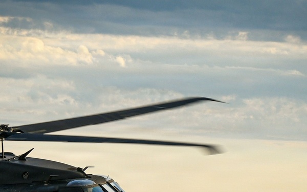 A military helicopter, identifiable as a UH-60 Black Hawk, is captured in flight against a partly cloudy sky at what appears to be dusk or golden hour. The helicopter is painted in dark olive/matte black military livery and has its side door open with crew members visible. The image is taken from another airborne platform, providing a close aerial perspective over a landscape of rolling hills and scattered settlements below.