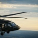 A military helicopter, identifiable as a UH-60 Black Hawk, is captured in flight against a partly cloudy sky at what appears to be dusk or golden hour. The helicopter is painted in dark olive/matte black military livery and has its side door open with crew members visible. The image is taken from another airborne platform, providing a close aerial perspective over a landscape of rolling hills and scattered settlements below.