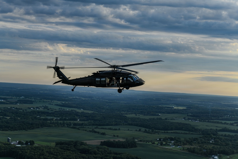 A dark military helicopter, consistent with a UH-60 Black Hawk, is captured in flight at altitude over a rural landscape of green fields and scattered tree lines. The image appears to be taken from another airborne platform during late afternoon or early evening, given the warm horizon light and overcast sky. The terrain below suggests a temperate agricultural region.