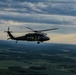 A dark military helicopter, consistent with a UH-60 Black Hawk, is captured in flight at altitude over a rural landscape of green fields and scattered tree lines. The image appears to be taken from another airborne platform during late afternoon or early evening, given the warm horizon light and overcast sky. The terrain below suggests a temperate agricultural region.