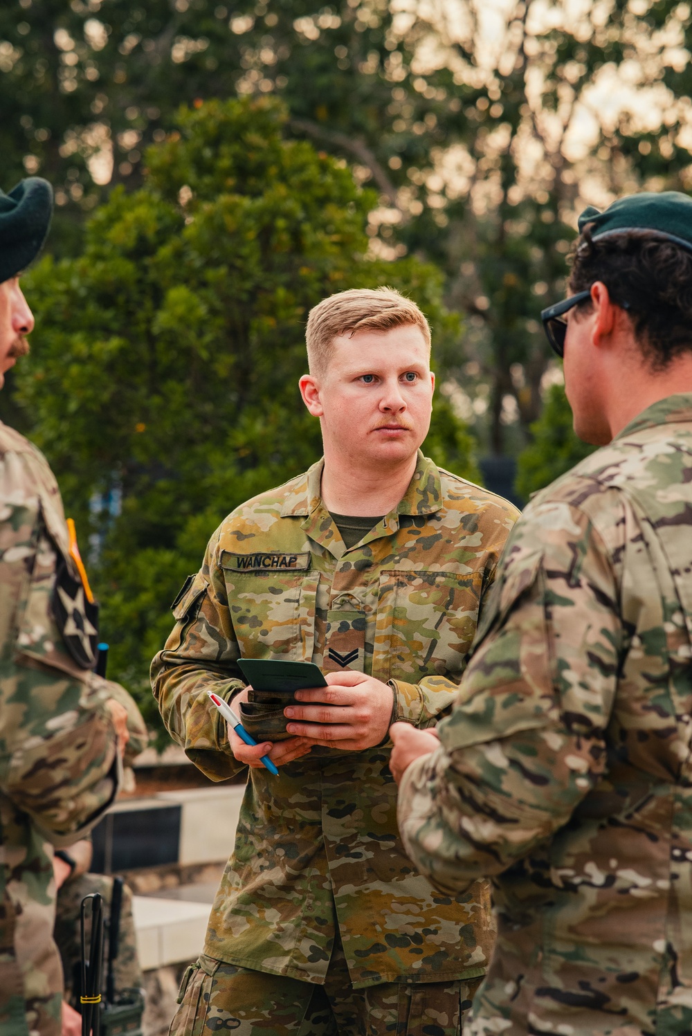 A military personnel member wearing Australian Army AUSCAM (AMCU) camouflage uniform with the name tape 'WANCHAP' and a Corporal rank insignia is holding a notebook and pen, appearing to be in a briefing or discussion. He is engaged in conversation with at least two other uniformed individuals in an outdoor setting with trees in the background. The lighting suggests either early morning or late afternoon.