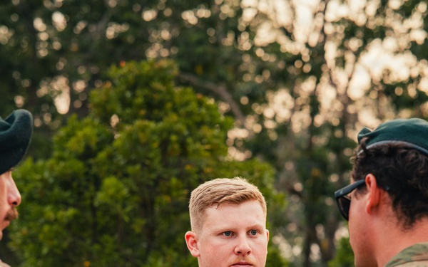 A military personnel member wearing Australian Army AUSCAM (AMCU) camouflage uniform with the name tape 'WANCHAP' and a Corporal rank insignia is holding a notebook and pen, appearing to be in a briefing or discussion. He is engaged in conversation with at least two other uniformed individuals in an outdoor setting with trees in the background. The lighting suggests either early morning or late afternoon.