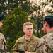 A military personnel member wearing Australian Army AUSCAM (AMCU) camouflage uniform with the name tape 'WANCHAP' and a Corporal rank insignia is holding a notebook and pen, appearing to be in a briefing or discussion. He is engaged in conversation with at least two other uniformed individuals in an outdoor setting with trees in the background. The lighting suggests either early morning or late afternoon.
