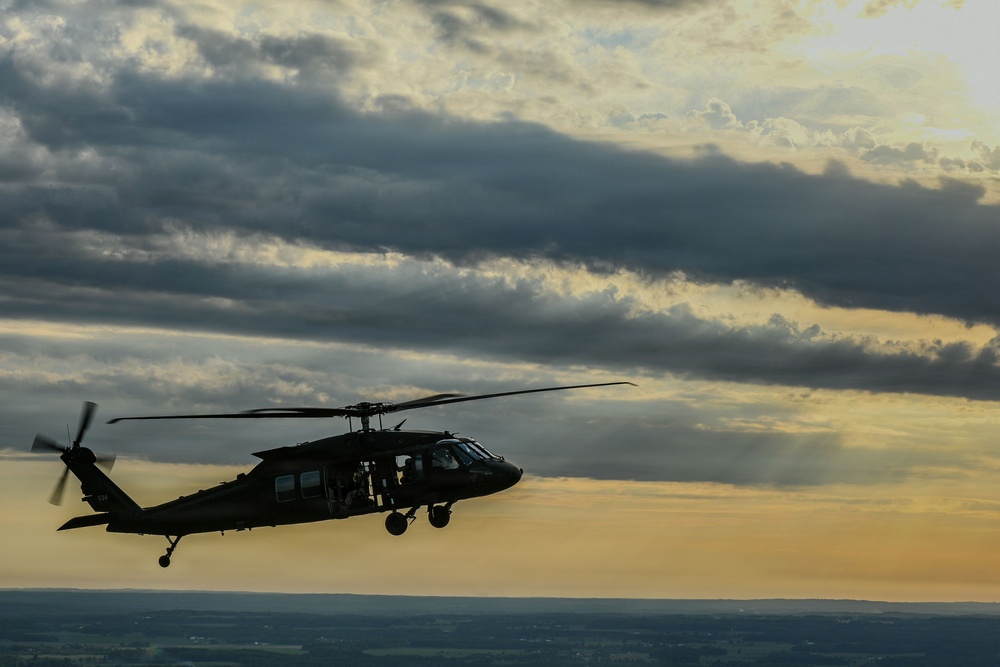 A military helicopter, consistent with a UH-60 Black Hawk design, is captured in silhouette flying against a dramatic sunset sky with layered clouds. The image is taken from an aerial perspective, suggesting it may have been photographed from another aircraft. Golden light breaks through the cloud cover on the horizon, creating a striking contrast with the dark helicopter silhouette.