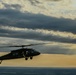 A military helicopter, consistent with a UH-60 Black Hawk design, is captured in silhouette flying against a dramatic sunset sky with layered clouds. The image is taken from an aerial perspective, suggesting it may have been photographed from another aircraft. Golden light breaks through the cloud cover on the horizon, creating a striking contrast with the dark helicopter silhouette.