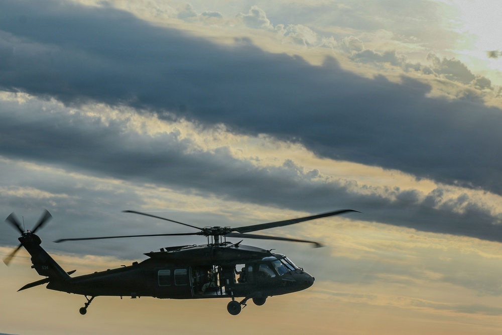 A military helicopter, consistent with a UH-60 Black Hawk design, is captured in flight against a dramatic cloudy sky at dusk or dawn. The aircraft is silhouetted in dark olive/black coloring with rotor blades in motion, suggesting active flight. The lighting conditions create a moody, atmospheric composition with warm golden tones breaking through the cloud cover.