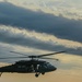 A military helicopter, consistent with a UH-60 Black Hawk design, is captured in flight against a dramatic cloudy sky at dusk or dawn. The aircraft is silhouetted in dark olive/black coloring with rotor blades in motion, suggesting active flight. The lighting conditions create a moody, atmospheric composition with warm golden tones breaking through the cloud cover.