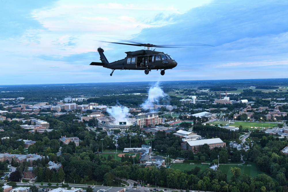 A black military helicopter, identifiable as a UH-60 Black Hawk, flies over what appears to be a large university campus with a football stadium visible below. White smoke or pyrotechnics are visible rising from the stadium area, suggesting a pre-game or ceremonial flyover event. The aerial perspective reveals an expansive campus with numerous buildings, athletic fields, and tree-lined grounds at dusk.
