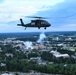 A black military helicopter, identifiable as a UH-60 Black Hawk, flies over what appears to be a large university campus with a football stadium visible below. White smoke or pyrotechnics are visible rising from the stadium area, suggesting a pre-game or ceremonial flyover event. The aerial perspective reveals an expansive campus with numerous buildings, athletic fields, and tree-lined grounds at dusk.