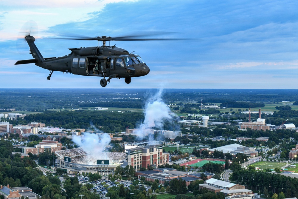 A military UH-60 Black Hawk helicopter flies over what appears to be a college football stadium during a game, with smoke or pyrotechnics visible rising from the stadium below. The aerial perspective reveals a large university campus surrounded by trees and suburban development. The scene suggests a pregame or halftime military flyover ceremony at a college football event.