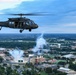 A military UH-60 Black Hawk helicopter flies over what appears to be a college football stadium during a game, with smoke or pyrotechnics visible rising from the stadium below. The aerial perspective reveals a large university campus surrounded by trees and suburban development. The scene suggests a pregame or halftime military flyover ceremony at a college football event.