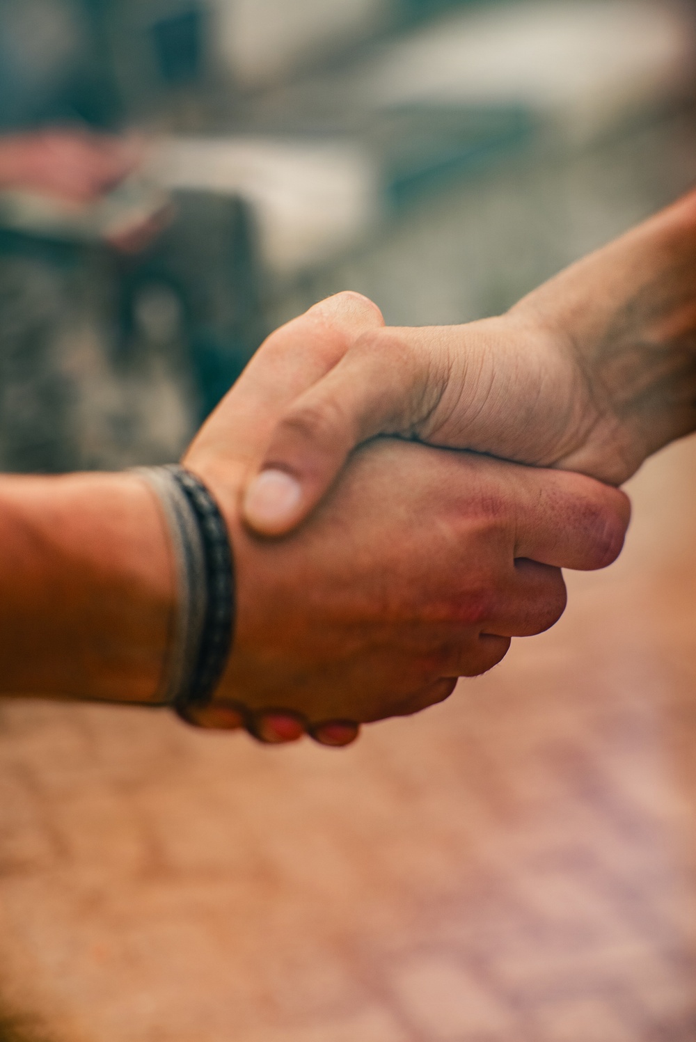 The image shows a close-up of two hands engaged in a firm handshake. One hand has a dark braided or woven bracelet visible on the wrist. The background is blurred with warm and cool tones, keeping the focus on the handshake.