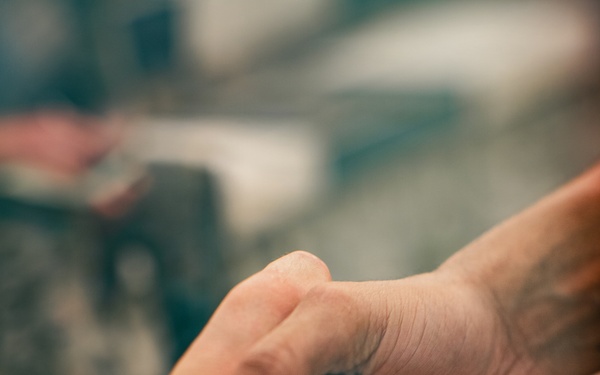 The image shows a close-up of two hands engaged in a firm handshake. One hand has a dark braided or woven bracelet visible on the wrist. The background is blurred with warm and cool tones, keeping the focus on the handshake.