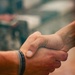 The image shows a close-up of two hands engaged in a firm handshake. One hand has a dark braided or woven bracelet visible on the wrist. The background is blurred with warm and cool tones, keeping the focus on the handshake.