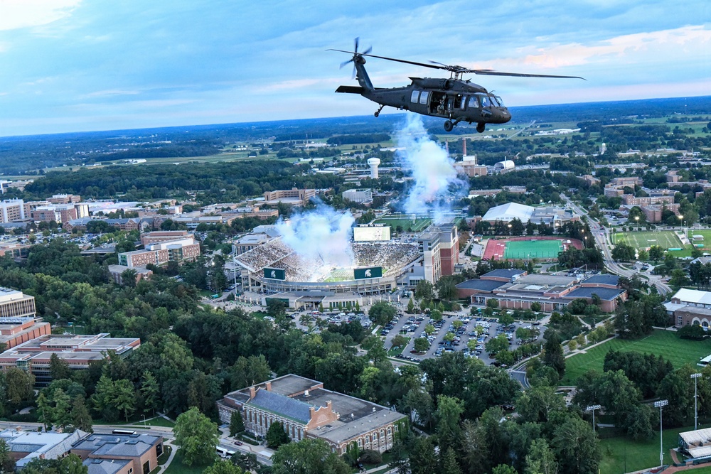 An aerial photograph captures a military UH-60 Black Hawk helicopter flying over a large university campus during what appears to be a football game event. Below, a packed stadium with green and white team colors is visible with pyrotechnic smoke rising from the field, consistent with a pre-game or halftime ceremony. The expansive campus, surrounded by dense tree cover and brick buildings, stretches to the horizon under a partly cloudy sky at dusk.
