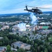 An aerial photograph captures a military UH-60 Black Hawk helicopter flying over a large university campus during what appears to be a football game event. Below, a packed stadium with green and white team colors is visible with pyrotechnic smoke rising from the field, consistent with a pre-game or halftime ceremony. The expansive campus, surrounded by dense tree cover and brick buildings, stretches to the horizon under a partly cloudy sky at dusk.