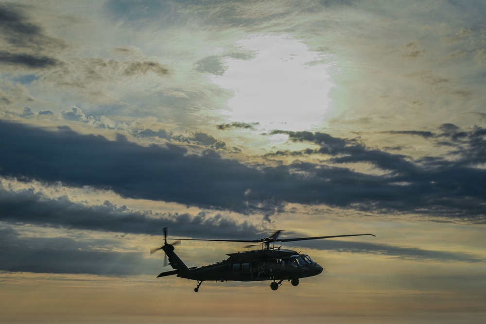 A military helicopter, consistent with a UH-60 Black Hawk silhouette, is captured in flight against a dramatic, cloud-filled sky at what appears to be dusk or dawn. The aircraft is rendered largely in dark silhouette due to backlighting from the bright sky. The image has a moody, atmospheric quality with layered clouds and warm golden tones near the horizon.
