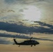 A military helicopter, consistent with a UH-60 Black Hawk silhouette, is captured in flight against a dramatic, cloud-filled sky at what appears to be dusk or dawn. The aircraft is rendered largely in dark silhouette due to backlighting from the bright sky. The image has a moody, atmospheric quality with layered clouds and warm golden tones near the horizon.