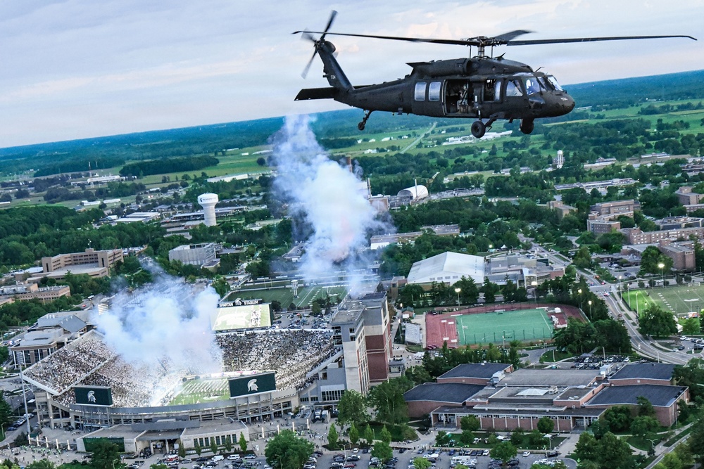 A U.S. Army UH-60 Black Hawk helicopter flies over what appears to be Spartan Stadium at Michigan State University during a pre-game or halftime ceremony. Pyrotechnic smoke effects are visible rising from the stadium, which is filled with spectators. The aerial perspective reveals the broader campus and surrounding East Lansing, Michigan landscape.