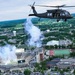 A U.S. Army UH-60 Black Hawk helicopter flies over what appears to be Spartan Stadium at Michigan State University during a pre-game or halftime ceremony. Pyrotechnic smoke effects are visible rising from the stadium, which is filled with spectators. The aerial perspective reveals the broader campus and surrounding East Lansing, Michigan landscape.