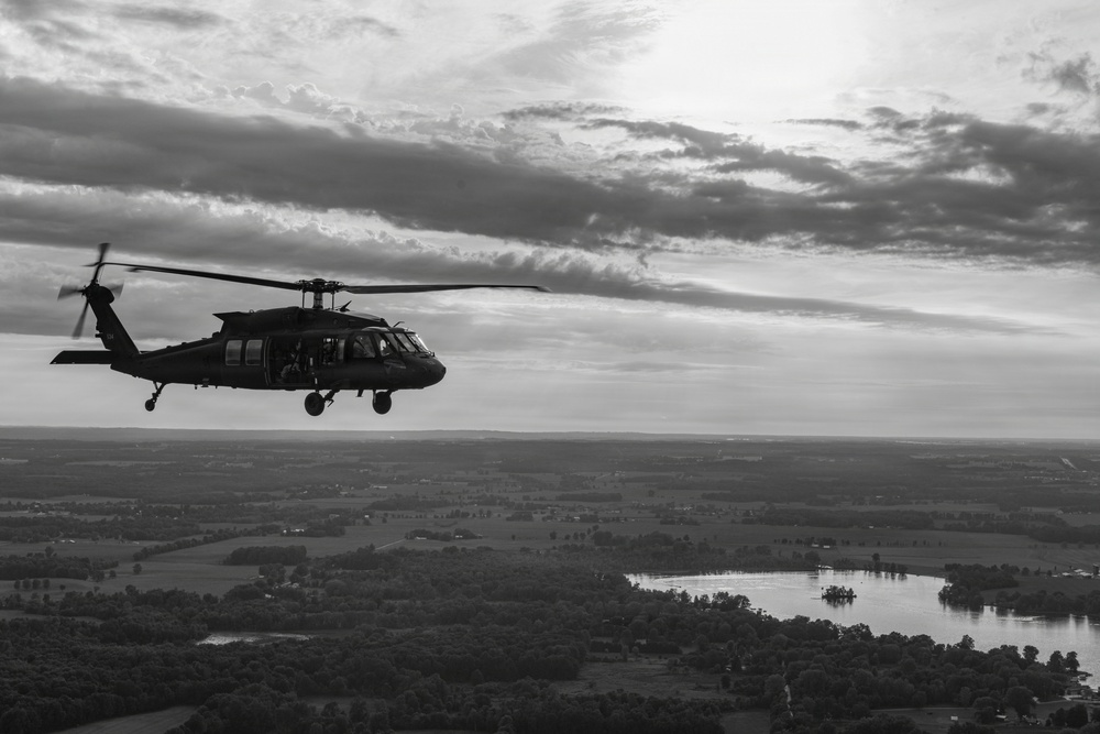 A black-and-white aerial photograph depicting a military helicopter in flight over a rural landscape. The helicopter is silhouetted against a dramatic, partly cloudy sky, with a river or lake visible in the background below. The image has a strong contrast and documentary-style composition.