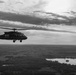 A black-and-white aerial photograph depicting a military helicopter in flight over a rural landscape. The helicopter is silhouetted against a dramatic, partly cloudy sky, with a river or lake visible in the background below. The image has a strong contrast and documentary-style composition.
