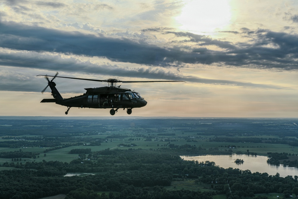 A military helicopter, consistent with a UH-60 Black Hawk, is photographed in flight at altitude during what appears to be dusk or dawn. The aircraft is silhouetted against a dramatic sky with scattered clouds and a low sun on the horizon. Below, a rural landscape with green fields, tree lines, and a visible lake or reservoir stretches to the horizon.