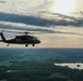 A military helicopter, consistent with a UH-60 Black Hawk, is photographed in flight at altitude during what appears to be dusk or dawn. The aircraft is silhouetted against a dramatic sky with scattered clouds and a low sun on the horizon. Below, a rural landscape with green fields, tree lines, and a visible lake or reservoir stretches to the horizon.
