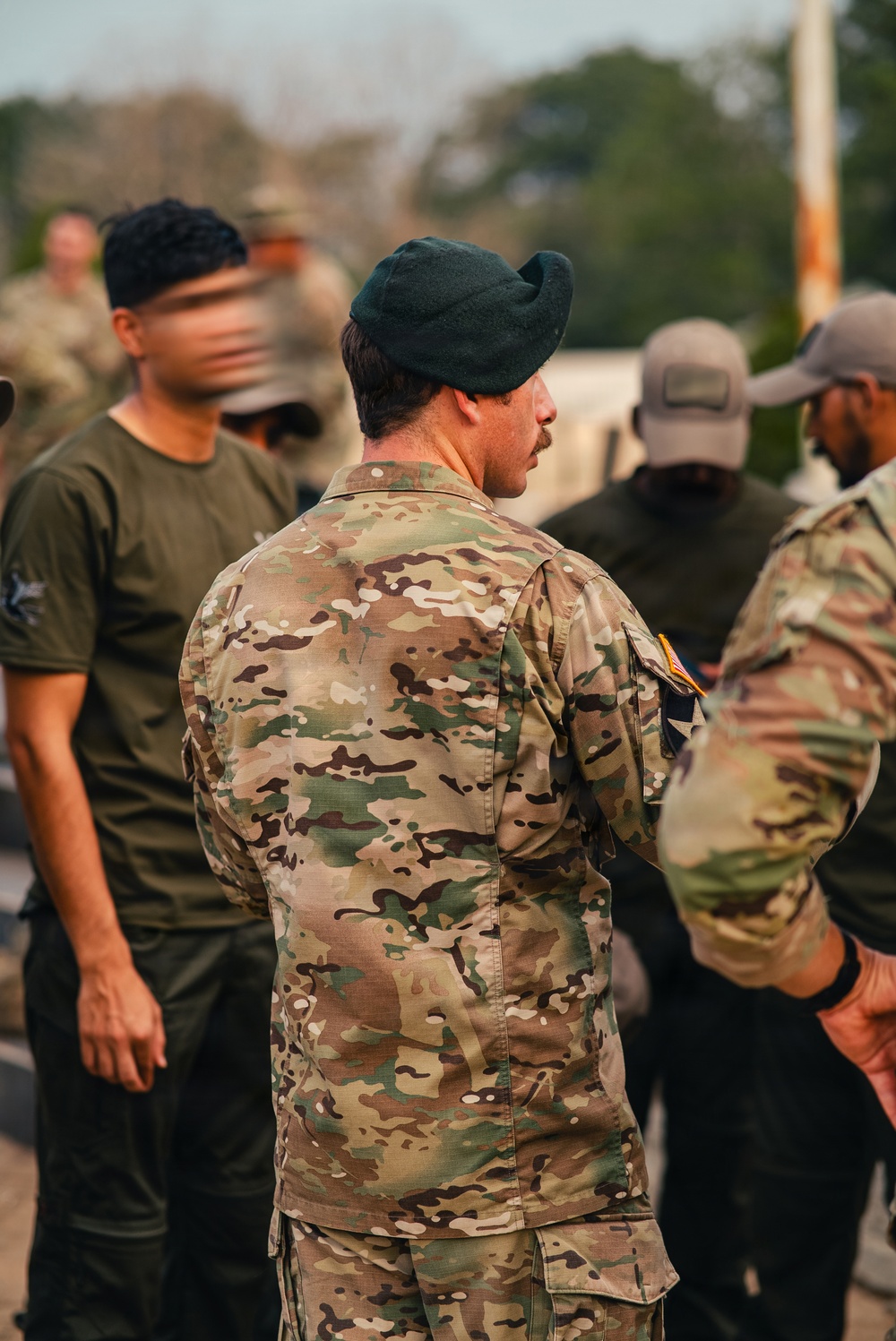 A group of military personnel gathered outdoors, with the central figure wearing OCP (Operational Camouflage Pattern) uniform and a dark green beret. Multiple individuals in various military and tactical attire are visible, suggesting a formation, briefing, or assembly event. The setting appears to be a military installation or training area with trees visible in the background.